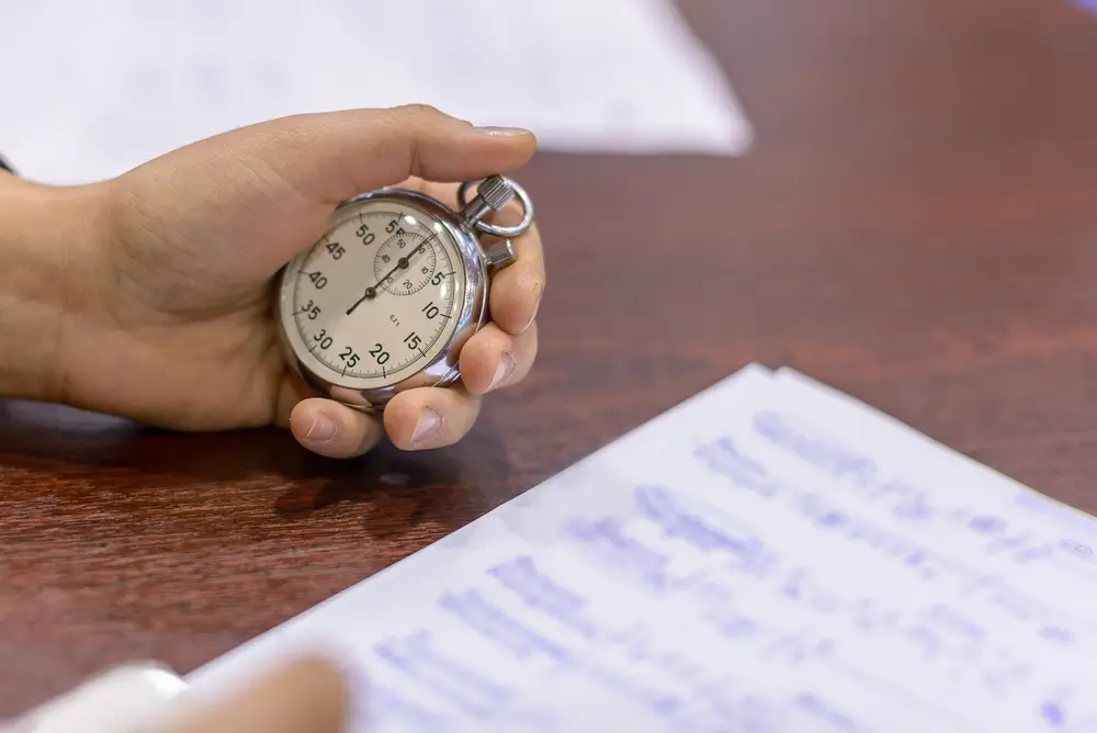 Mano de un opositor usando un cronómetro analógico para medir el tiempo mientras practica un examen tipo test.
