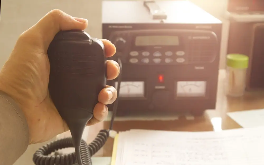 Mano sosteniendo un micrófono de radio policial frente a una emisora de base sobre un escritorio con documentos.