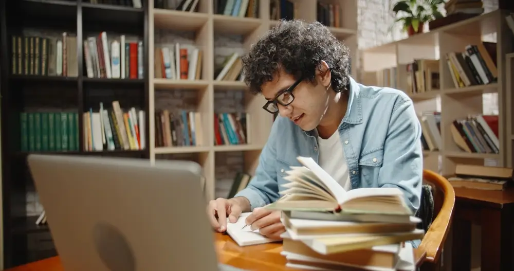 Estudiante joven concentrado preparando una oposición con libros y ordenador en una biblioteca.
