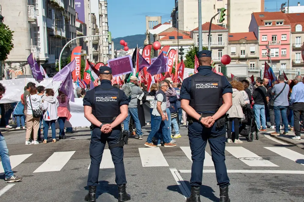 Inspector de la Policía Nacional con uniforme operativo y chaleco de espaldas vigilando una manifestación urbana.