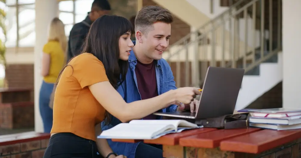 Dos jóvenes estudiantes consultan un portátil y libros para preparar oposiciones en una zona de estudio al aire libre.