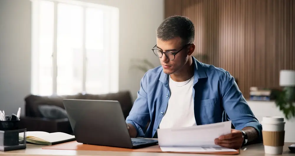 Estudiante usando un ordenador portátil y revisando documentos para su oposición a la Policía Nacional.